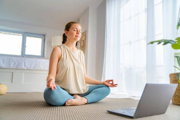 Mädchen sitzt im Schneidersitz auf dem Boden ihrer Wohnung vor ihrem Laptop in einem Zoom Call und führt eine Meditationsübung bei der Soulwork Academy aus